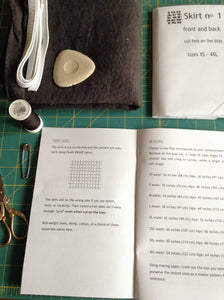 open instruction book. folded sewing pattern, pile of fabric, elastic and tailor's chalk, spool of thread, safety pin and small scissors