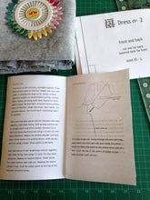 open instruction booklet with folded pattern. An arrangement of ruler, folded fabric and colorful pins