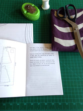 open instruction booklet and folded sewing pattern. Arrangement of folded fabric, large scissors, pin cushion and a spool of thread