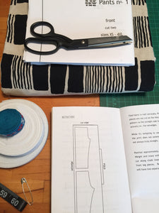 Open instruction book with scissors on top of a folded sewing pattern and fabric. Rolled elastic, fabric trim, safety pin, measuring tape